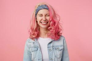 feliz fofa sorridente senhora com Rosa cabelo e tatuado mãos, esperando para surpresa com fechadas olhos, amplamente sorridente, em pé sobre Rosa fundo, vestindo uma branco camiseta e jeans jaqueta. foto