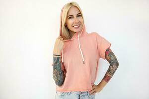 alegre jovem bonita Loiras fêmea com tatuagens mostrando dela branco perfeito dentes enquanto sorridente alegremente para Câmera, posando sobre branco fundo dentro Rosa camiseta foto