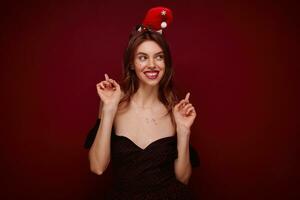 estúdio tiro do jovem elegante alegre senhora com tarde Maquiagem levantando acima indicadores e sorridente Felizmente, desfrutando x-mas tema festa mascarada enquanto posando sobre clarete fundo foto