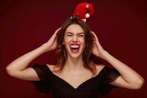 muito feliz lindo jovem morena fêmea vestido dentro festivo roupas vestindo santa chapéu e segurando mãos em dela cabeça, rindo alegremente e demonstrando dela branco perfeito dentes sobre clarete fundo foto