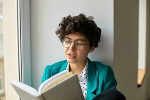 retrato do encantador jovem encaracolado morena mulher com curto na moda corte de cabelo guardando livro dentro elevado mãos enquanto sentado sobre peitoril da janela em brilhante dia dentro à moda elegante vestem foto