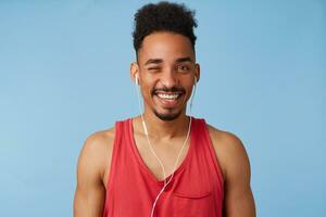 foto do jovem africano americano feliz homem detém, escuta legal músicas, desgasta dentro uma vermelho camisa, aparência às Câmera e piscadelas, carrinhos sobre azul fundo.