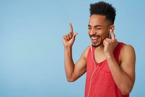 feliz africano americano atraente homem dentro uma vermelho jérsei escuta legal música, detém fone de ouvido com esquerda mão, fecha dele olhos, alto canta e danças, carrinhos sobre azul fundo. foto