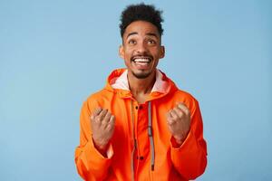 jovem atraente africano americano Sombrio esfolado cara desgasta dentro laranja chuva casaco, sente muito feliz, sorrisos amplamente, Veja acima e aperta dele punhos carrinhos sobre azul fundo. foto