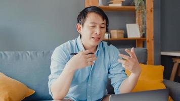 jovem empresário asiático usa fones de ouvido usando laptop e fala com colegas sobre o plano de videochamada enquanto trabalha em casa na sala de estar. auto-isolamento, distanciamento social, quarentena para prevenção ambiciosa.