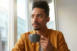 retrato do jovem bonito Sombrio esfolado duvidando barista, bebidas aromático café a partir de uma cinzento cam e pensativamente parece ausente, tentando para gosto a gosto do grão. foto