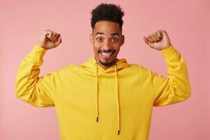 jovem alegre africano americano homem dentro amarelo capuz, levantando dele punhos acima, dizer eu sou legal' e carrinhos sobre Rosa fundo e sorridente amplamente. foto