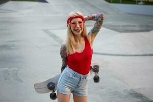 agradável Loiras senhora com tatuado braços vestido dentro uma vermelho camiseta e jeans shorts, com uma tricotado bandana em dela cabeça, dentro vermelho copos, segurando uma Longboard dentro dela mãos e amplamente sorridente. foto