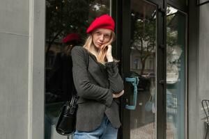 encantador jovem elegante Loiras mulher dentro elegante roupas inclinado dela cabeça em elevado mão e olhando calmamente às Câmera com guardada lábios, posando sobre cafeteria exterior foto