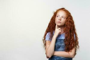 foto do pensando fofa sardas pequeno menina com gengibre cabelo, parece sério e dúvida, toques queixo, parece longe sobre branco fundo com cópia de espaço em a esquerda lado.