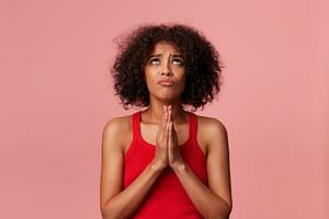 retrato do jovem beleza africano americano menina com encaracolado Sombrio cabelo vestindo dentro uma vermelho camiseta. haha acima, mantém Palmeiras junto, satisfeito para misericórdia. isolado sobre Rosa fundo. foto