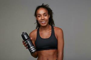 retrato do jovem desportivo alegre Sombrio esfolado mulher com encaracolado Castanho cabelo segurando ginástica garrafa dentro elevado mão e olhando alegremente às Câmera com sincero sorriso, posando sobre cinzento fundo foto