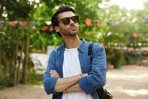 ao ar livre retrato do bonita jovem barbudo masculino dentro oculos de sol posando sobre verde parque com cruzado braços em dele peito, olhando a parte, de lado com sério face foto