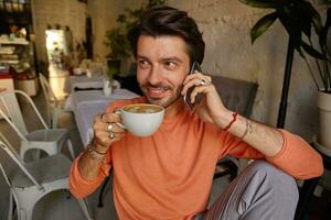feliz atraente masculino com barba bebendo café enquanto tendo conversação em Móvel telefone, posando sobre cafeteria interior, ser dentro agradável humor foto