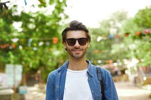 goog olhando jovem Sombrio cabelos masculino com barba vestindo casual roupas e oculos de sol, em pé sobre verde parque em ensolarado caloroso dia, positivo emoções conceito foto