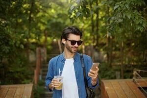 jovem positivo bonito masculino com barba caminhando através cidade parque em ensolarado caloroso dia, verificação mensagens em dele Móvel telefone e bebendo suco foto