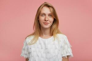 encantador jovem ruiva mulher com natural Maquiagem olhando positivamente às Câmera e sorridente suavemente enquanto posando sobre Rosa fundo dentro branco elegante camiseta foto