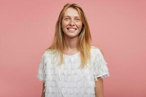 retrato do jovem feliz adorável ruiva mulher com casual Penteado mostrando dela branco perfeito dentes enquanto sorridente alegremente, em pé contra Rosa fundo foto