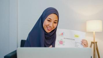 senhora muçulmana asiática usa hijab usando computador laptop conversa com um colega sobre o plano de uma reunião de videochamada enquanto trabalha remotamente em casa à noite na sala de estar. distanciamento social, quarentena para o vírus corona.