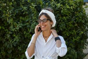 retrato do bonita jovem fêmea olhando a parte, de lado com encantador sorrir enquanto tendo agradável conversação em dela telefone, vestindo branco na moda roupas e vintage oculos de sol foto