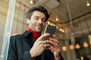 fechar-se do elevado do homem mãos com argolas guardando Móvel telefone enquanto posando sobre cidade cafeteria fundo, mensagens de texto mensagem para amigos e sorridente levemente enquanto olhando positivamente em tela foto