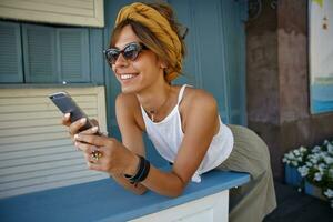 retrato do alegre jovem senhora com casual Penteado vestindo branco topo e Oliva saia, inclinado em bancada e olhando a parte, de lado com encantador sorriso, guardando Smartphone dentro mãos foto