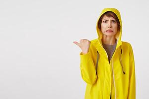 atraente menina dentro uma amarelo capa de chuva com uma com nojo expressão em dela face quer para desenhar seu atenção para a cópia de espaço em a esquerda apontando com dele dedo, em pé sobre branco muro. foto