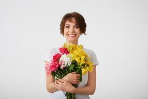 retrato do feliz sorridente agradável jovem curto cabelos mulher dentro branco em branco camiseta, segurando uma ramalhete do colorida flores com fechadas olhos, em pé sobre branco fundo. foto