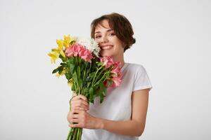 feliz fofa jovem curto cabelos mulher dentro branco em branco camiseta, amplamente sorridente, em pé sobre branco fundo, segurando uma ramalhete do colorida flores, desfrutando a cheiro. foto