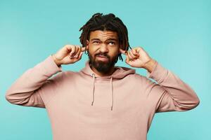 descontente jovem bonito Sombrio cabelos masculino com barba fazendo careta dele face e cobertura orelhas enquanto tentando para evitar alto sons, em pé sobre azul fundo foto