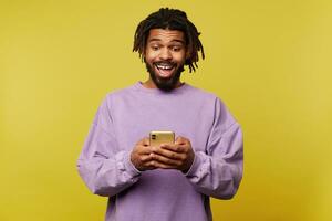 alegre jovem atraente Sombrio esfolado morena cara com barba segurando Móvel telefone e olhando excitadamente em tela com Largo boca aberto, posando sobre amarelo fundo foto