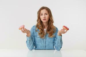 retrato do confusão jovem bonita mulher com grandes loiro ondulado cabelo, sentado às a mesa. difícil escolha entre dois rosquinhas que 1 sabores Melhor foto