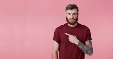 descontente jovem bonito vermelho barbudo homem dentro vermelho camisa, quer para desenhar seu atenção para cópia de espaço em a esquerda lado, franze a testa face dentro desagrado, carrinhos sobre Rosa fundo. foto