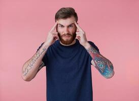 retrato do jovem tatuado vermelho barbudo homem dentro em branco camiseta, sente dor de cabeça e toques templos, carrinhos sobre Rosa fundo e franzindo a testa. foto