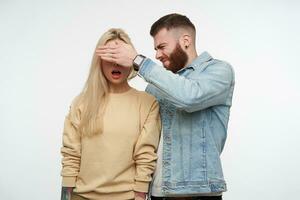 confuso jovem barbudo homem dentro jeans casaco franzindo a testa dele face e cobertura olhos do dele espantado grandes cabelos Loiras namorada dentro bege suéter, isolado sobre branco fundo foto