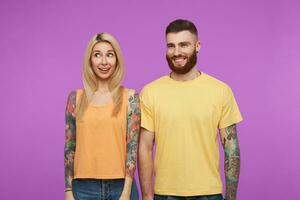 estúdio foto do jovem atraente tatuado casal vestido dentro laranja casual vestem guardando mãos ao longo seus corpos enquanto olhando alegremente aparte, isolado sobre roxa fundo