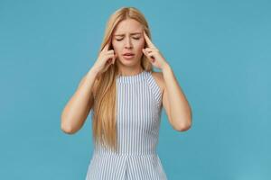 cansado jovem Loiras mulher com grandes cabelo guardando indicadores em templos, fechamento olhos Porque do dor de cabeça, posando sobre azul fundo dentro listrado verão vestir foto