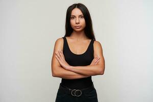 retrato do lindo jovem Sombrio cabelos mulher com casual Maquiagem dobrando mãos em dela peito enquanto olhando calmamente às Câmera, vestido dentro Preto roupas enquanto em pé sobre branco fundo foto