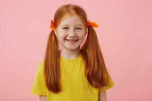 retrato do feliz petite sardas ruivo menina com dois caudas, amplamente sorridente e parece bonitinho, desgasta dentro amarelo camiseta, carrinhos sobre Rosa fundo. foto