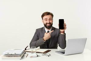 feliz sorridente atraente barbudo homem de negocios, topo Gerente sentado às Área de Trabalho dentro escritório, olhando Câmera, vestido dentro a caro terno com uma gravata, apontando com dedo para dele Smartphone. foto