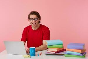 foto do feliz jovem aluna dentro óculos desgasta dentro vermelho camiseta, homem senta de a mesa e trabalhando com computador portátil e livros, amplamente sorrisos, parece alegre, isolado sobre Rosa fundo.