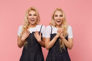 agitado jovem adorável grandes cabelos Loiras mulheres rindo alegremente enquanto olhando às Câmera e levantando emocionalmente mãos, isolado sobre Rosa fundo foto