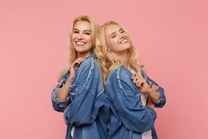 alegre jovem atraente grandes cabelos Loiras irmãs olhando de bom grado às Câmera com encantador sorrisos enquanto em pé costas para costas contra Rosa fundo foto