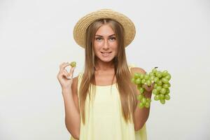 retrato do encantador jovem grandes cabelos Loiras senhora com casual Penteado vestindo velejador chapéu enquanto posando branco fundo, morder lábio inferior e segurando grupo do uvas foto