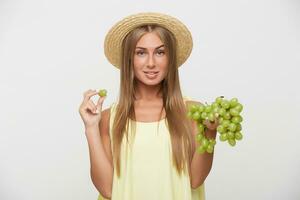 estúdio foto do lindo jovem Loiras mulher com natural Maquiagem levantando sobrancelha enquanto olhando às Câmera e segurando grupo do uvas enquanto em pé sobre branco fundo