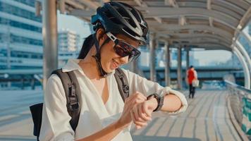empresária asiática com mochila bicicleta sorrindo look smartwatch na rua da cidade ir trabalhar no escritório. garota de esportes usa seu aplicativo de relógio para rastreamento de fitness. comutar para trabalhar, viajante de negócios na cidade.