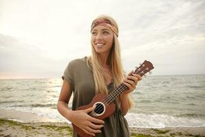 alegre jovem adorável grandes cabelos Loiras mulher com natural maekup sorridente amplamente enquanto olhando alegremente aparte, posando sobre de praia fundo com ukulele foto