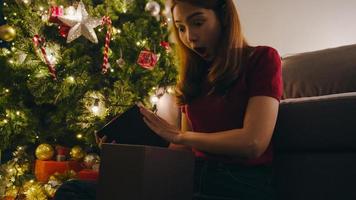 jovem asiática se divertindo, abrindo a caixa de presente de x'mas perto da árvore de Natal, decorada com enfeites na sala de estar em casa. feliz noite de Natal e feliz ano novo festival de férias. foto