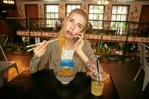 horizontal tiro do bonita jovem Loiras mulher com tatuagens tendo lince dentro moderno cafeteria, vestindo bege camisa e branco camiseta, tendo entediante conversação em dela telefone foto