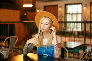 horizontal tiro do atraente tatuado jovem mulher dentro casual roupas sentado às mesa dentro cidade cafeteria e bebendo fresco suco, guardando Móvel telefone dentro mão e fazer ligar foto